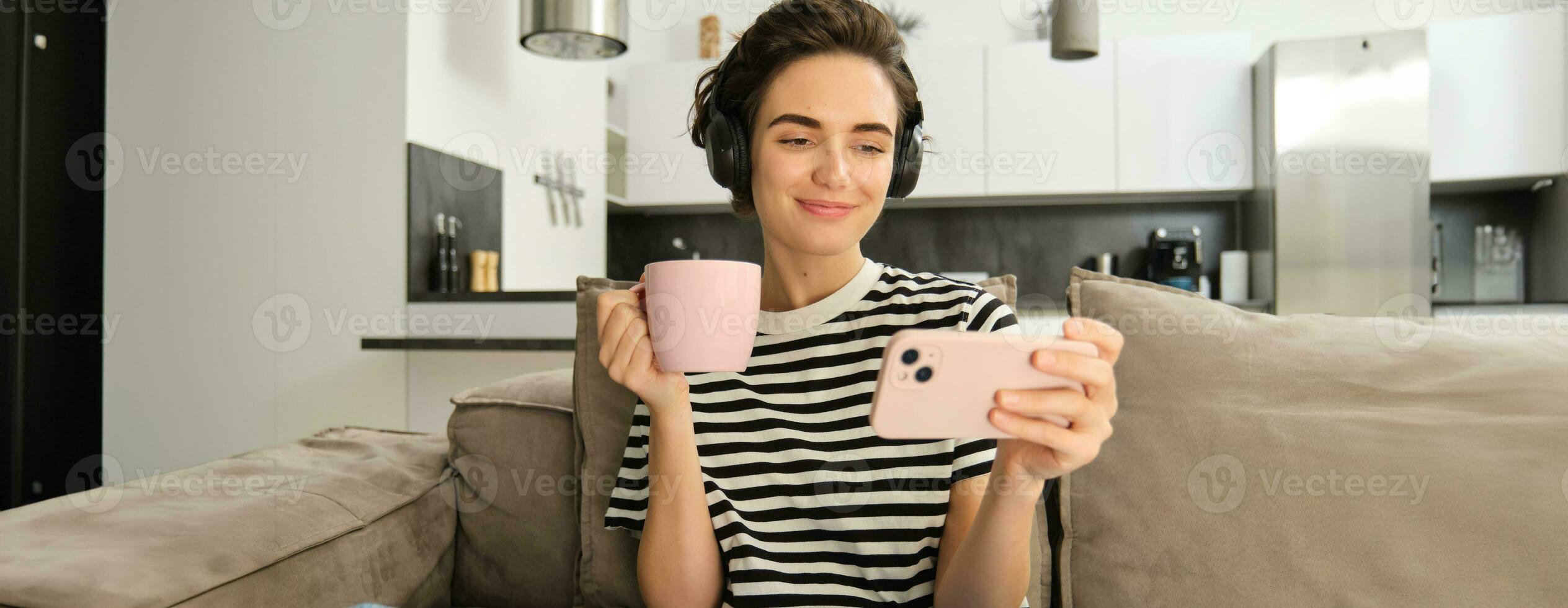 portret van vrouw in haar leven kamer, zittend Aan sofa en aan het kijken videos Aan mobiel telefoon, vervelend koptelefoon, drinken thee of koffie in roze kop foto