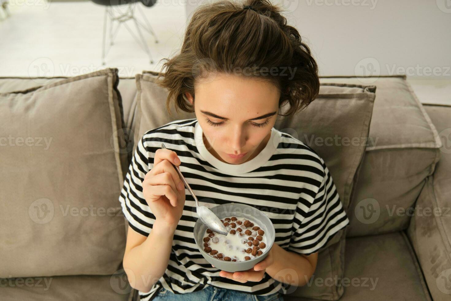 bovenste hoek schot van Vrolijk, schattig jong vrouw Aan bank, aan het eten kom van granen met melk en lachend, genieten van haar ontbijt foto