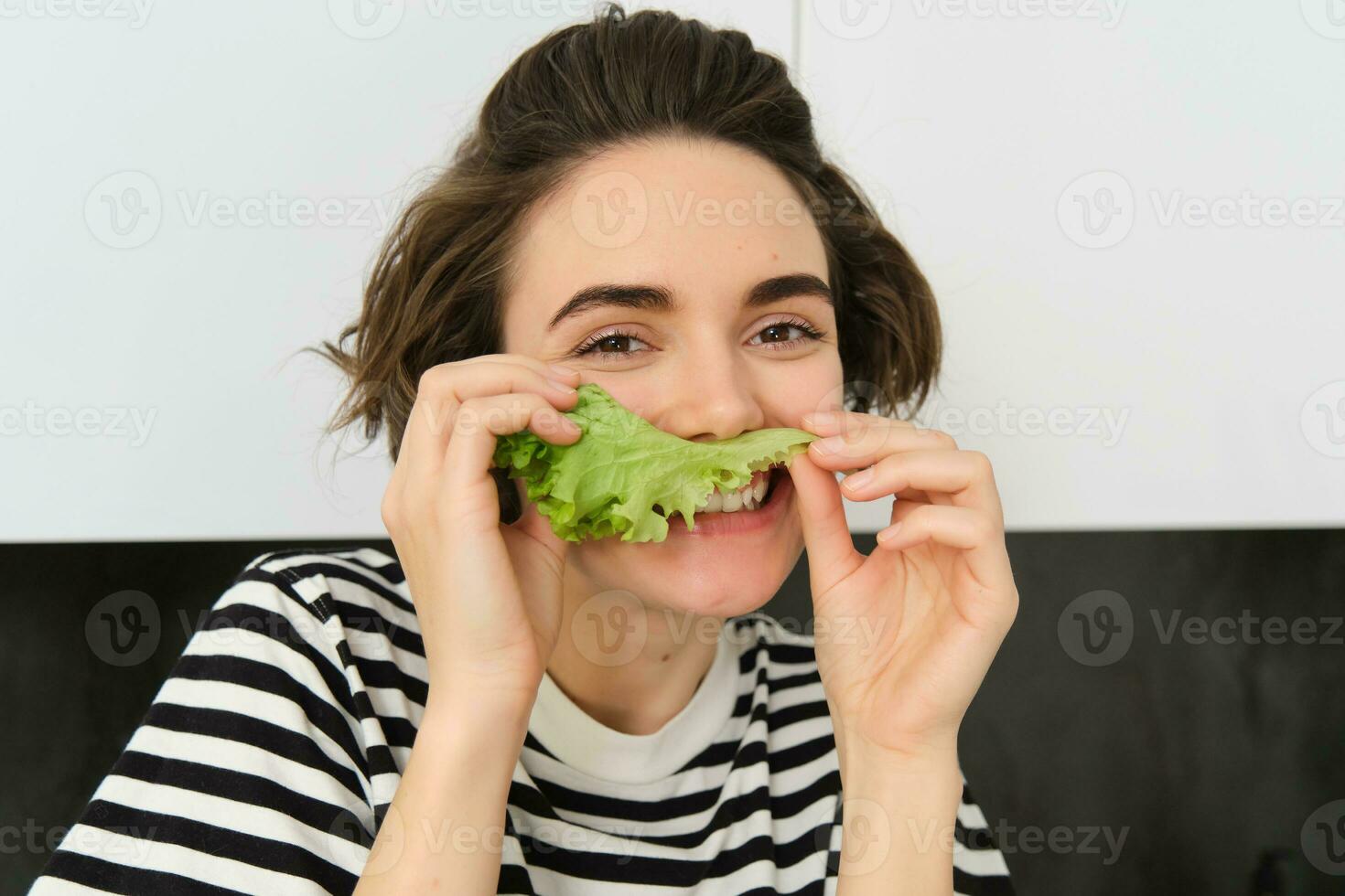 dichtbij omhoog portret van jong vrouw, vegetarisch meisje, houdt aan het eten groenten, poseren met sla blad en lachend, poseren in de keuken. concept van gezond voedsel en eetpatroon foto