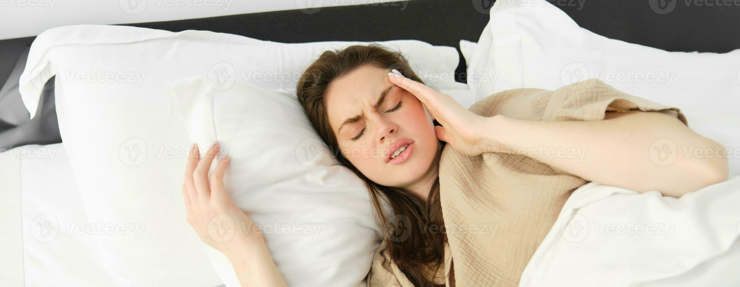 dichtbij omhoog portret van ziek vrouw aan het liegen in haar bed en gevoel onwel, aanraken hoofd, heeft hoofdpijn, op zoek gefrustreerd van migraine, vervelend pyjama foto