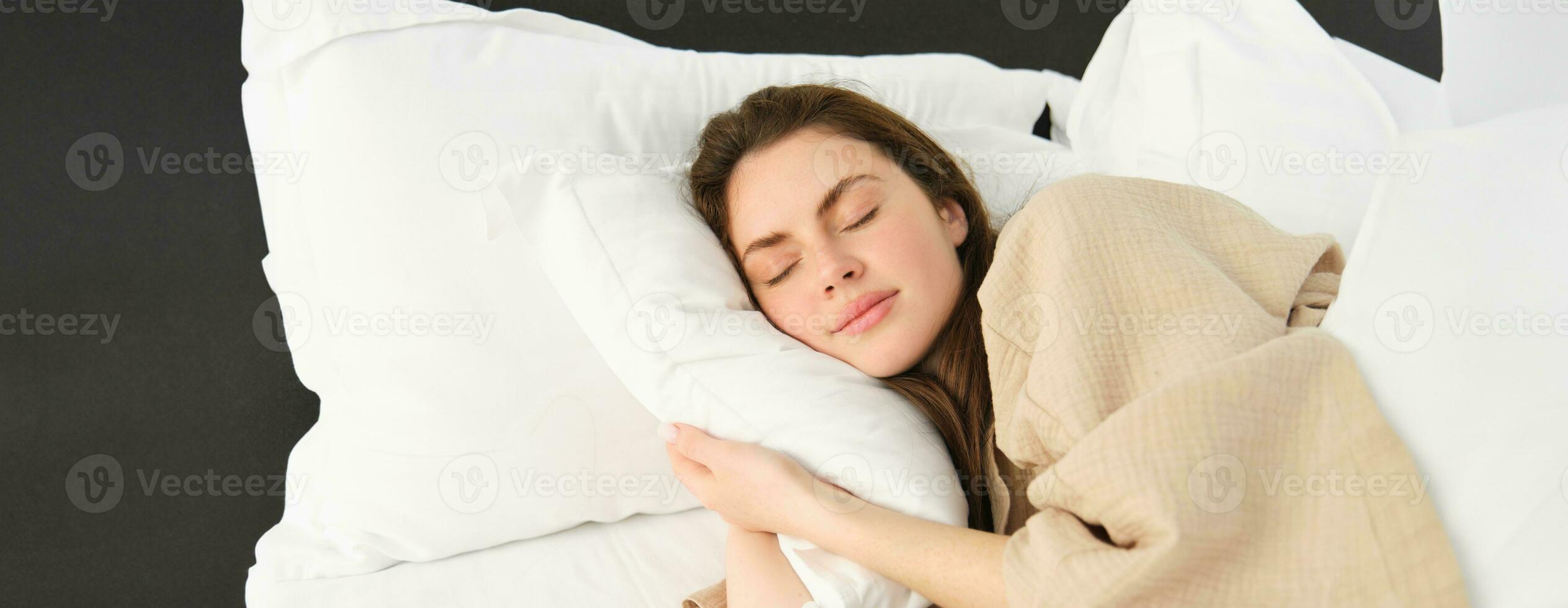 portret van Vrolijk, mooi jong vrouw resting in bed, vervelend pyjama, knuffelen haar hoofdkussen en glimlachen terwijl slapen foto