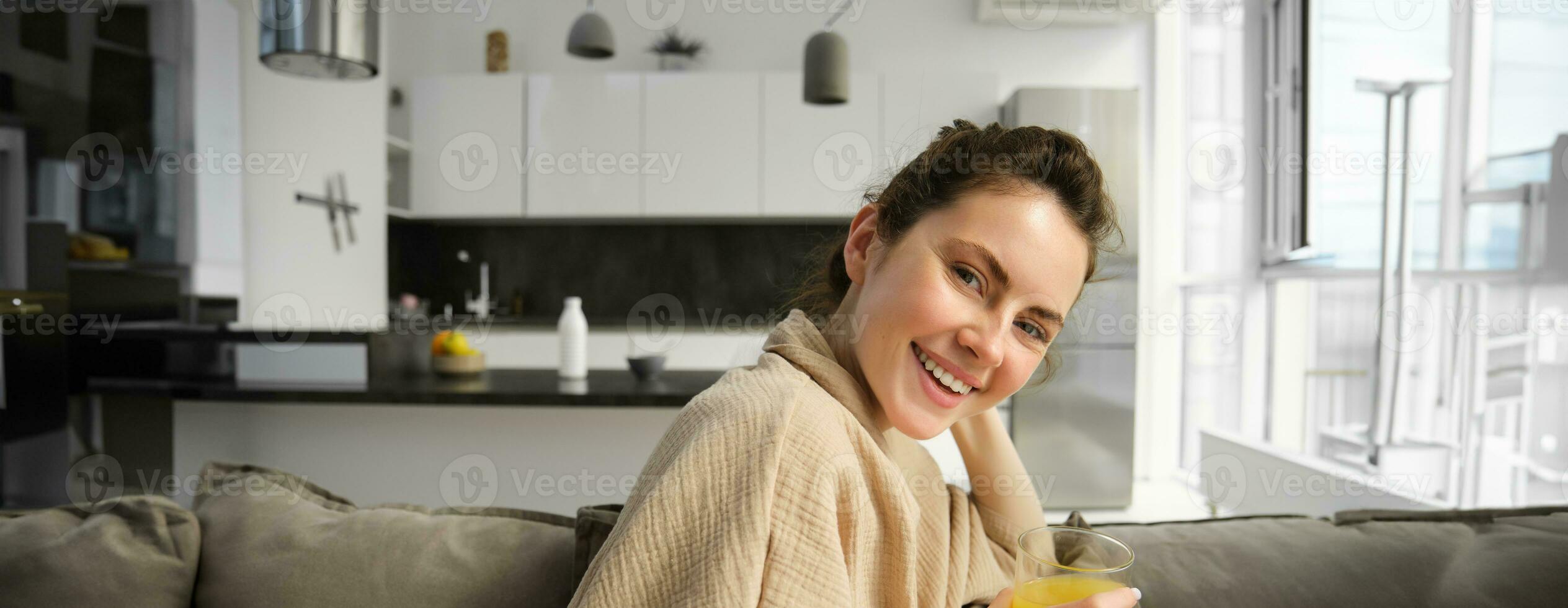 Dames welzijn en levensstijl. mooi jong vrouw Aan bank, drinken oranje sap, ontspannende Bij huis Aan bank, resting in de ochtend- in leven kamer foto