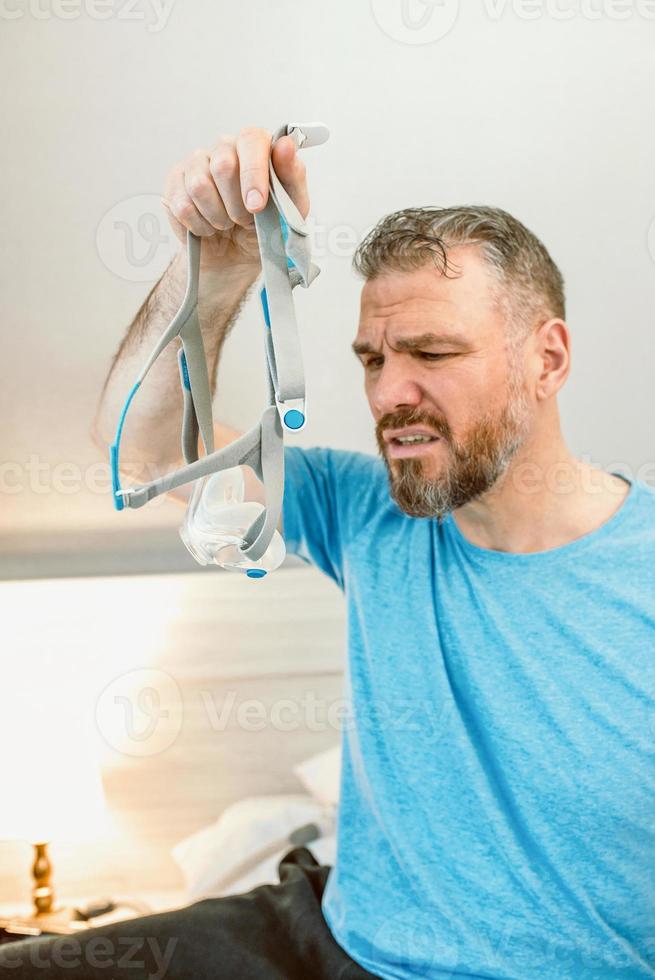 ongelukkige geschokte man met chronische ademhalingsproblemen verrast door het gebruik van een cpap-machine die op het bed in de slaapkamer zit. gezondheidszorg, cpap, obstructieve slaapapneu-therapie, snurkconcept foto