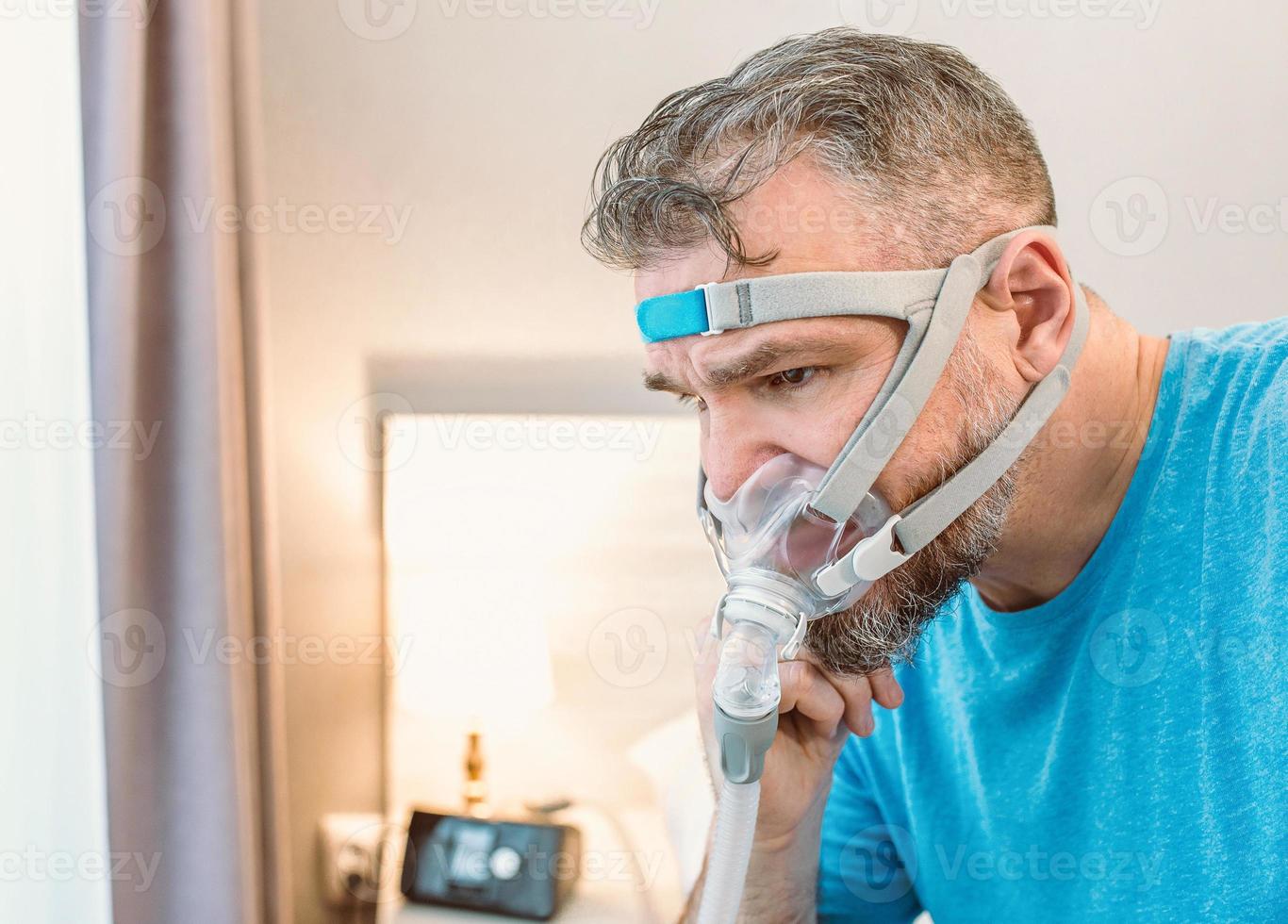 ongelukkige geschokte man met chronische ademhalingsproblemen verrast door het gebruik van een cpap-machine die op het bed in de slaapkamer zit. gezondheidszorg, cpap, obstructieve slaapapneu-therapie, snurkconcept foto