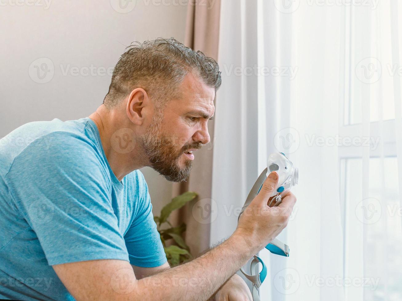 ongelukkige geschokte man met chronische ademhalingsproblemen verrast door het gebruik van een cpap-machine die op het bed in de slaapkamer zit. gezondheidszorg, cpap, obstructieve slaapapneu-therapie, snurkconcept foto