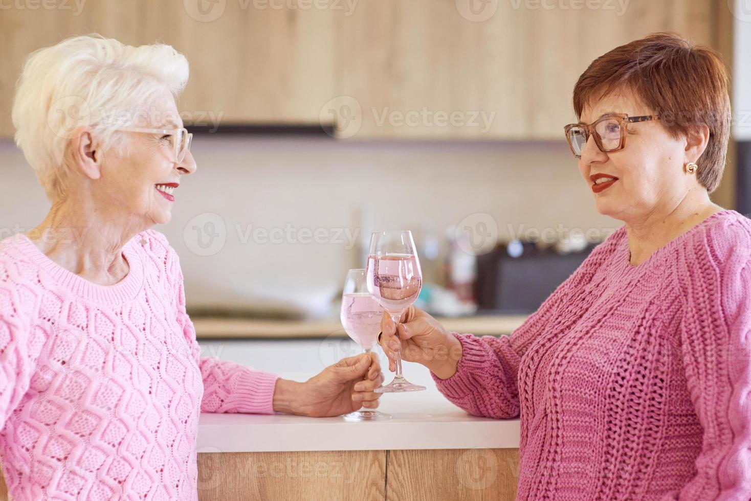 twee stijlvolle senior vrouwen in roze truien drinken rose wijn bij moderne keuken roddelen. vriendschap, praten, roddels, evenement, relaties, nieuws, familieconcept foto