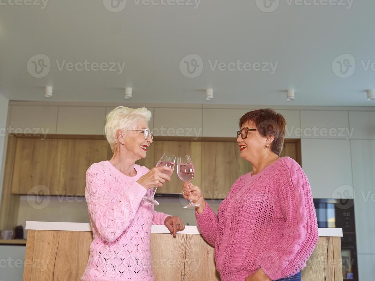 twee stijlvolle senior vrouwen in roze truien drinken rose wijn bij moderne keuken roddelen. vriendschap, praten, roddels, evenement, relaties, nieuws, familieconcept foto