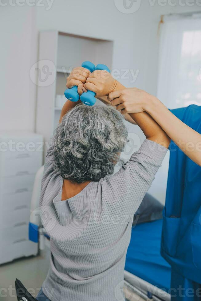 oud vrouw opleiding met fysiotherapeut gebruik makend van halters Bij huis. therapeut assisteren senior vrouw met opdrachten in verpleging huis. ouderen geduldig gebruik makend van halters met uitgestrekt armen. foto