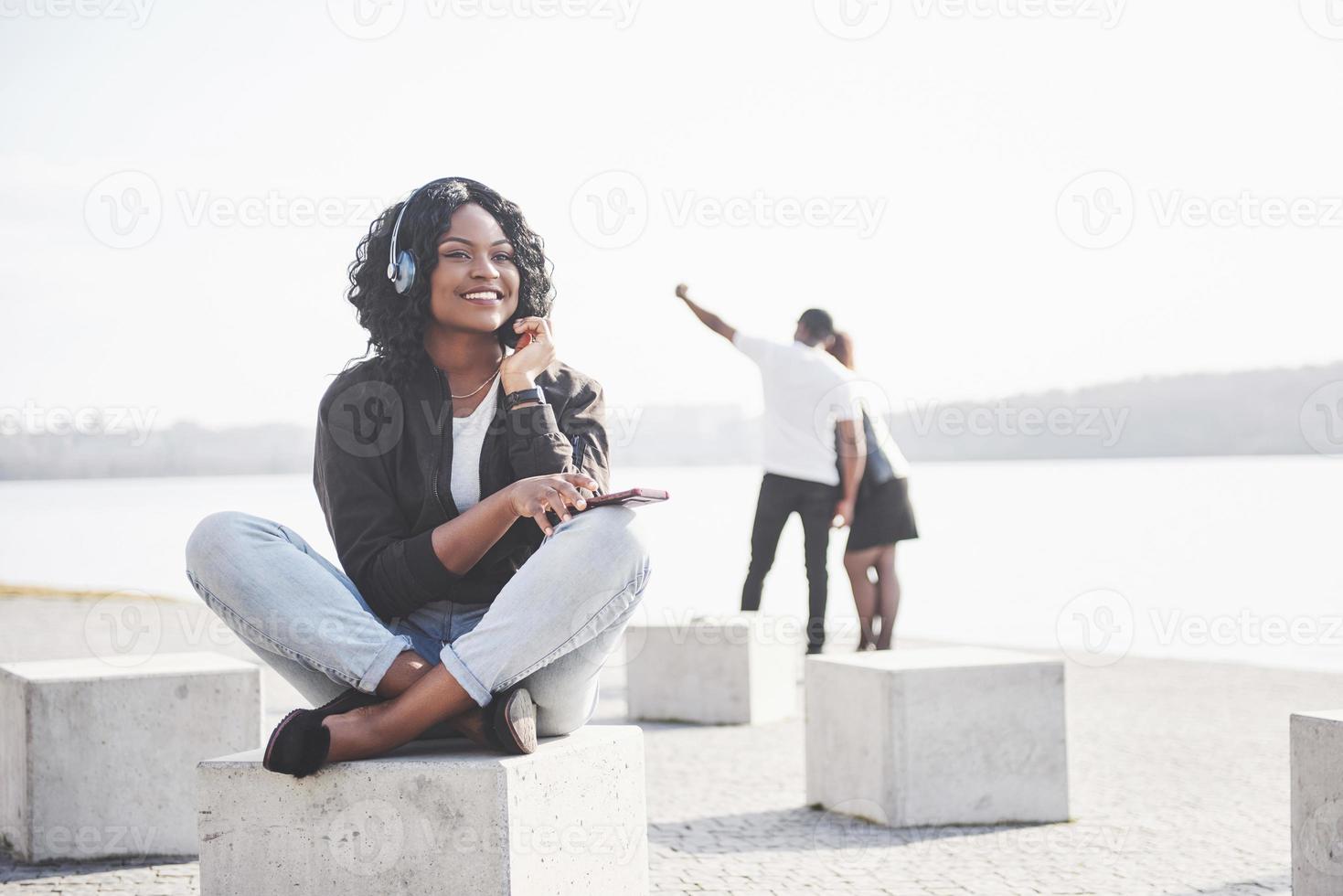 portret van een mooi jong mooi Afrikaans Amerikaans meisje zittend op het strand of het meer en luisterend naar muziek in haar koptelefoon foto