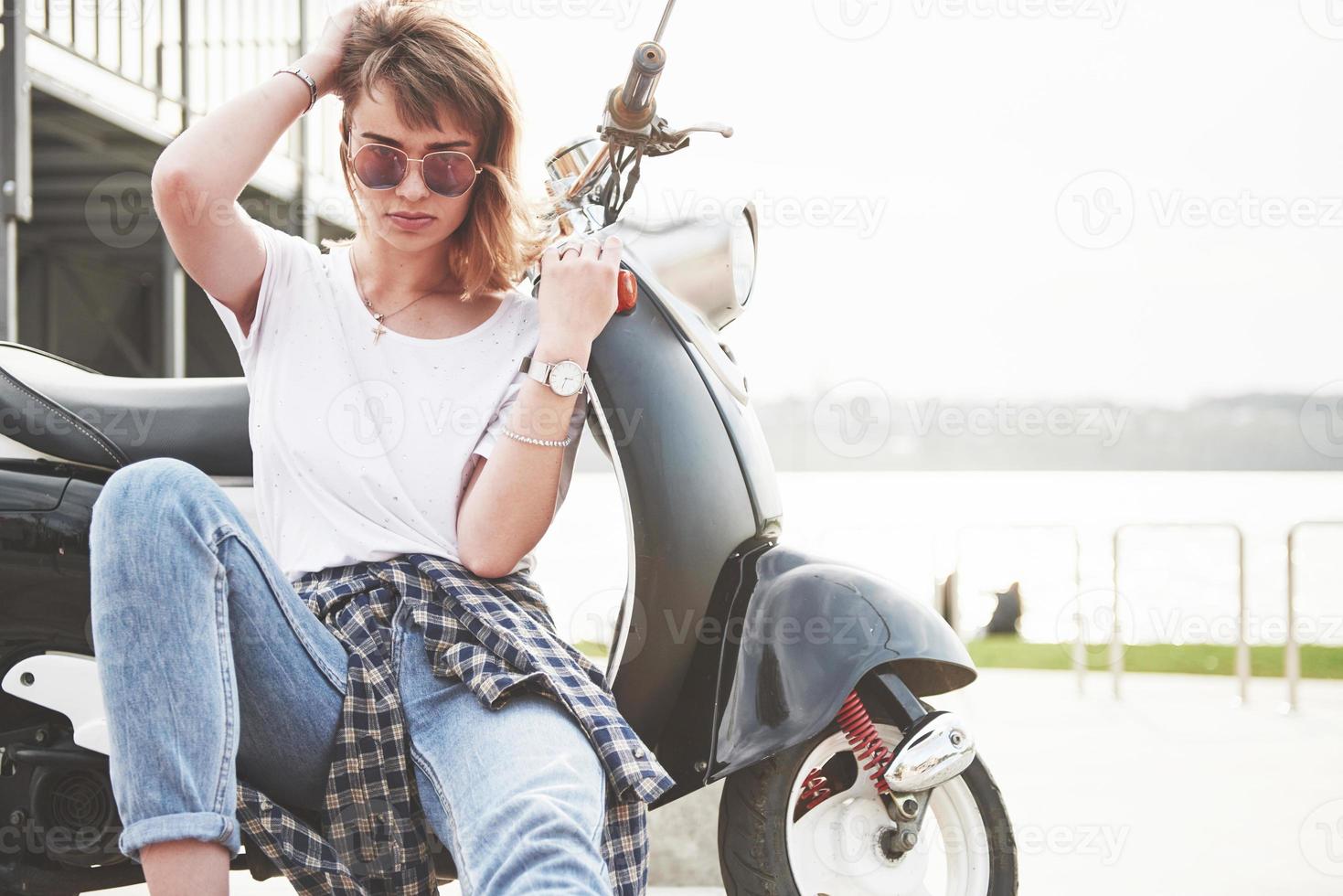 portret van een mooi meisje hipster zittend op een zwarte retro scooter, glimlachend poseren en genieten van de warme lentezon. foto