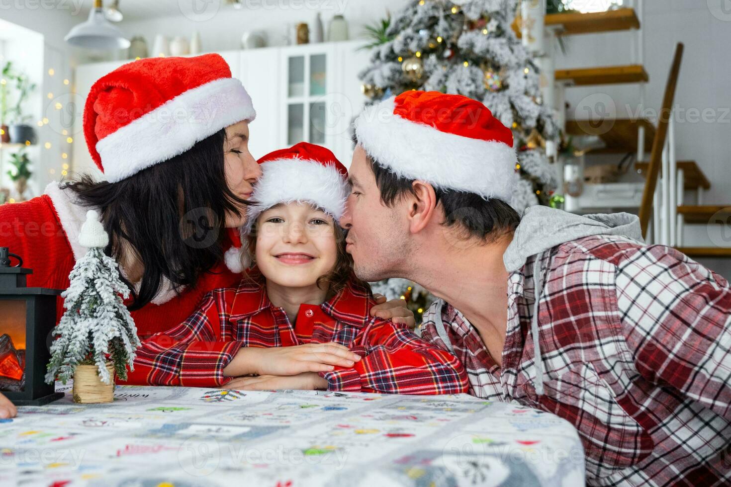 gelukkig traditioneel familie van pa, mama, dochter in feestelijk de kerstman hoeden in een wit keuken met een Kerstmis boom en decor. nieuw jaar, familie waarden. foto