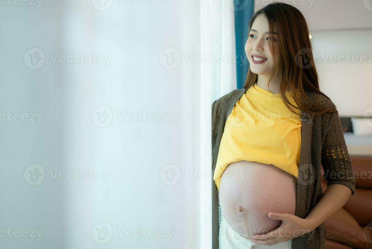 mooi zwanger vrouw staand in de buurt venster Bij huis en Holding haar buik foto