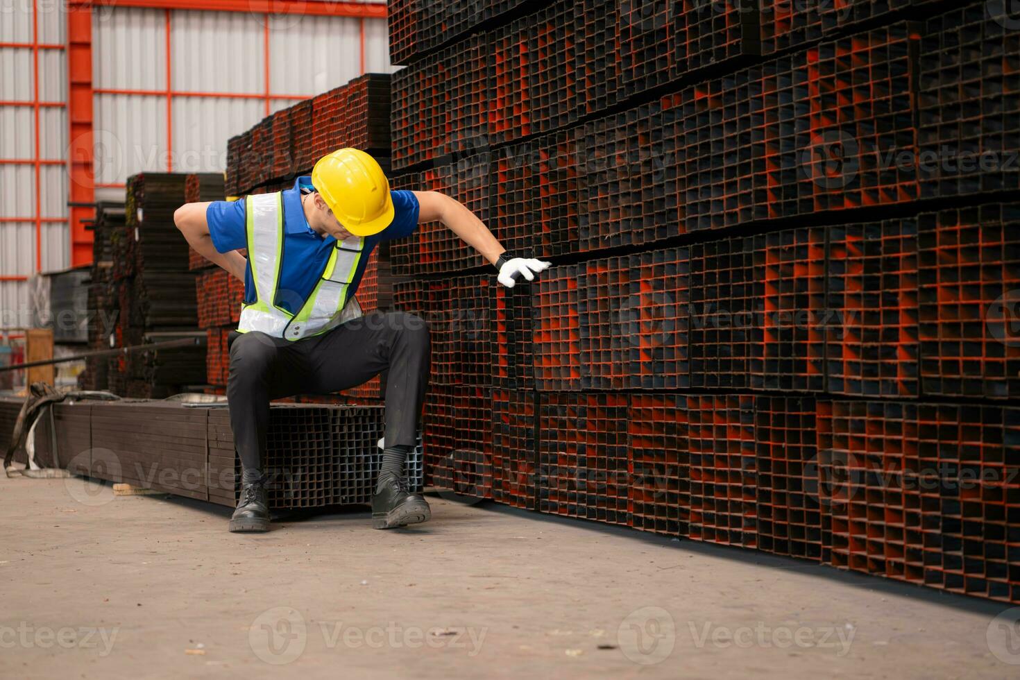 portret van een mannetje arbeider vervelend een veiligheid hesje en helm zittend Aan een staal pallet ten gevolge naar terug pijn van werken in een fabriek hijs- zwaar dingen. foto