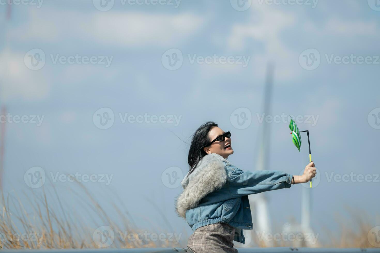 jong prachtig brunette vrouw met lang haar- vervelend een blauw jasje, zwart zonnebril, en Holding een miniatuur windmolen foto