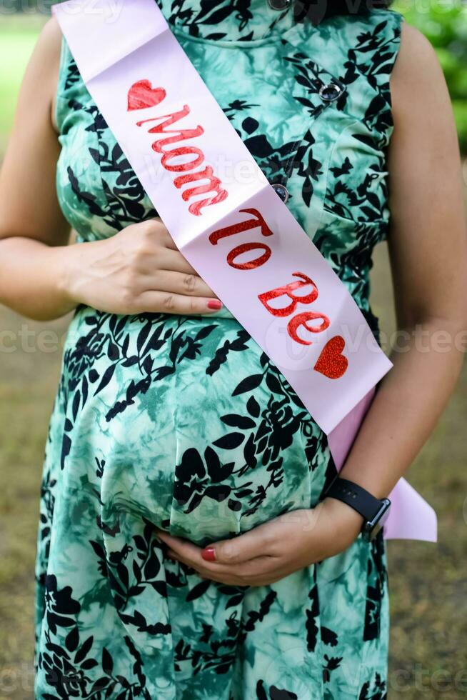 een zwanger Indisch dame poses voor buitenshuis zwangerschap schieten en handen Aan buik, Indisch zwanger vrouw zet haar hand- Aan haar maag met een moederschap jurk Bij maatschappij park, zwanger buiten moederschap schieten foto