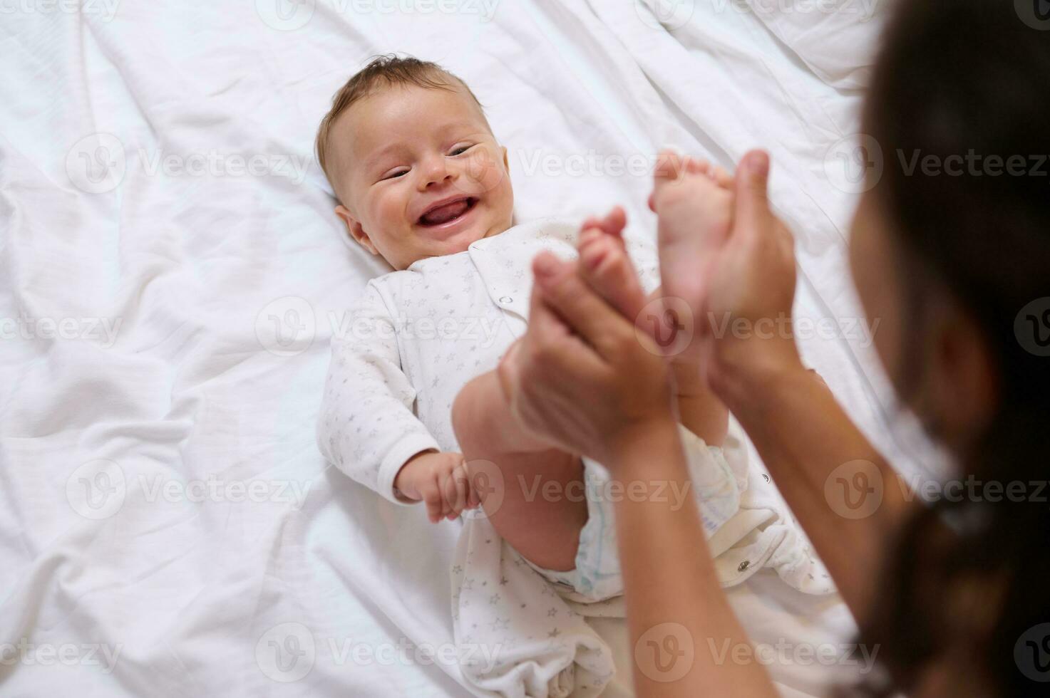 mooi pasgeboren baby glimlacht op zoek Bij zijn liefhebbend moeder, terwijl ze beroertes en kusjes zijn weinig voeten en klein tenen foto