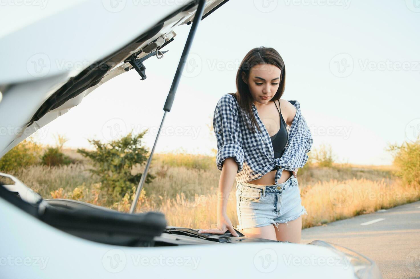 aantrekkelijk slank jong meisje in zomer shorts en overhemd reparaties een gebroken auto. een mooi vrouw staat in de buurt verheven auto kap. foto