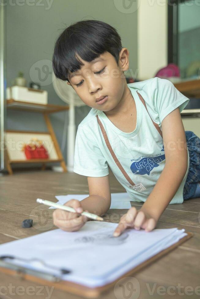 dichtbij omhoog portret van Aziatisch jongen met zwart pony, zwart ogen met een glimlachen gezicht vervelend een licht groen en aan het liegen Aan de verdieping van zijn huis tekening een afbeelding van een meisje met vlechtjes. onderwijs concept. foto