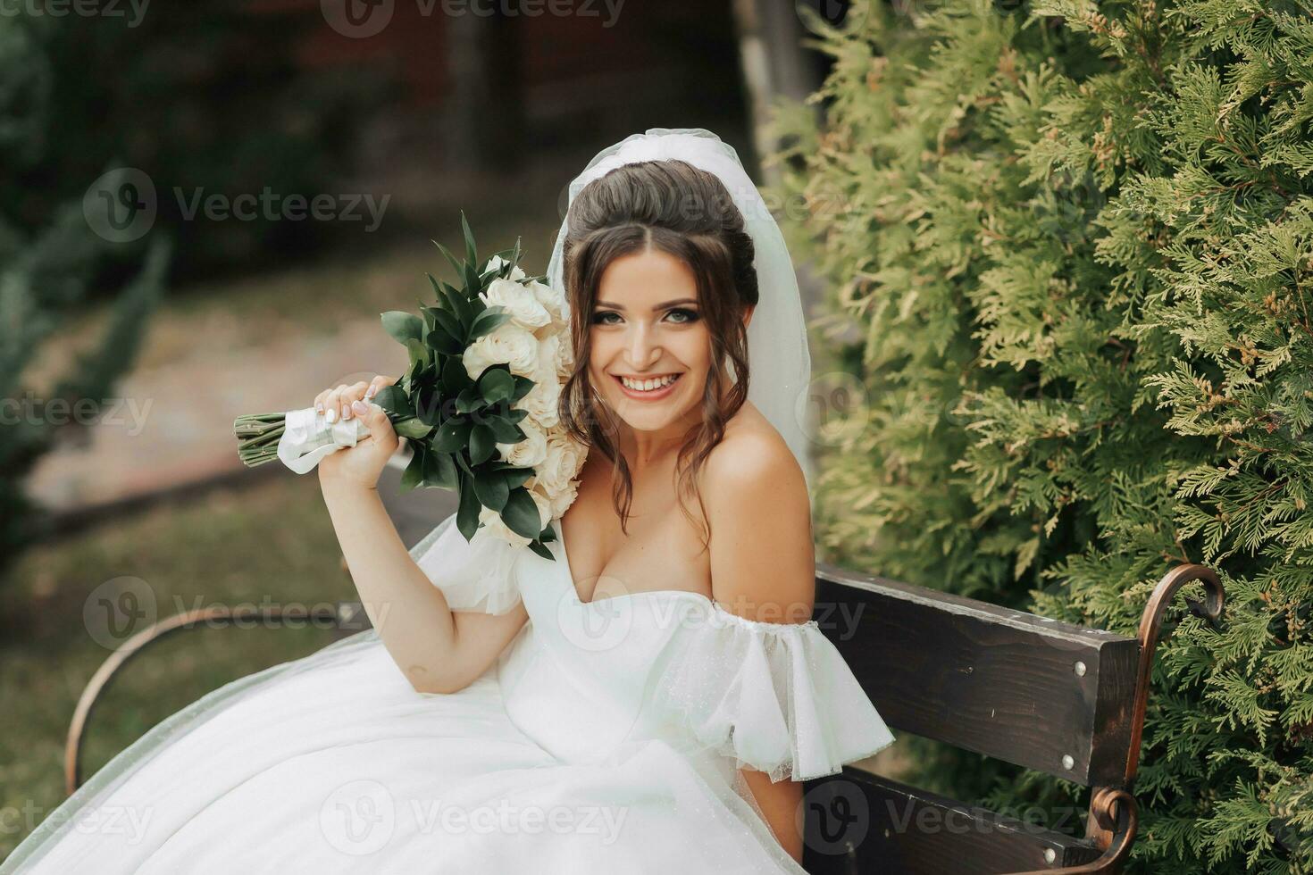 portret van de bruid in natuur. een brunette bruid in een wit lang jurk, Holding een boeket van wit rozen, poseert, op zoek in de lens, glimlachen oprecht. Aan de achtergrond van de boom. gekruld haar. foto