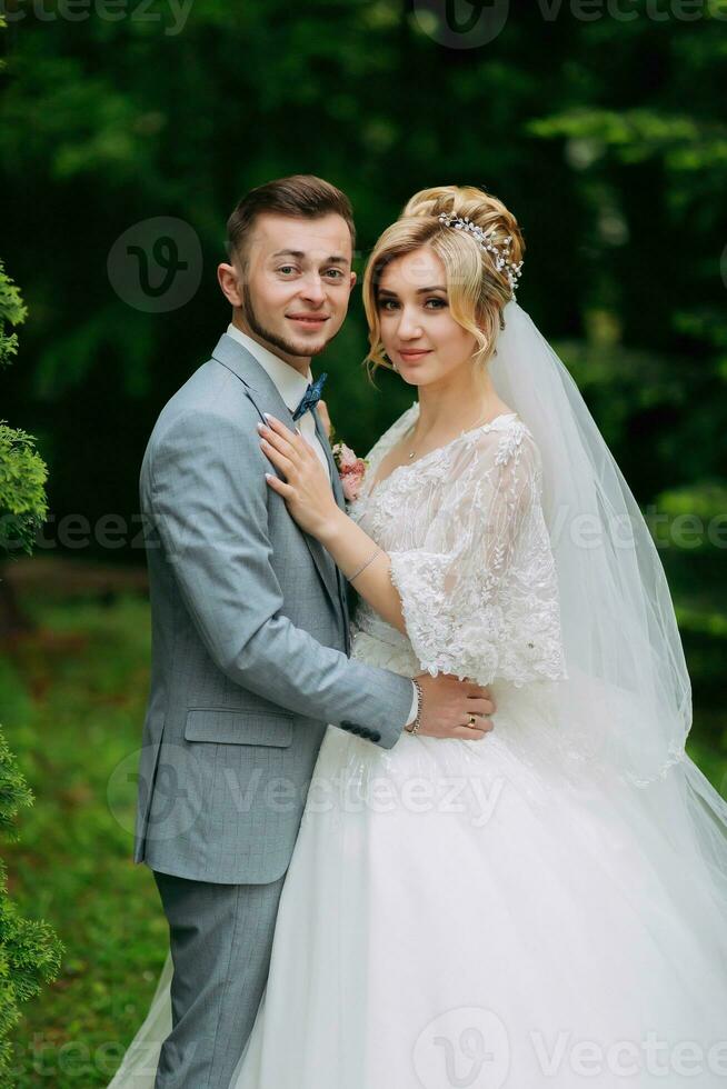 portret van de bruid en bruidegom staand tegen de achtergrond van groen bomen, knuffelen en op zoek in de lens. elegant bruidegom. mode en stijl. mooi bruid foto