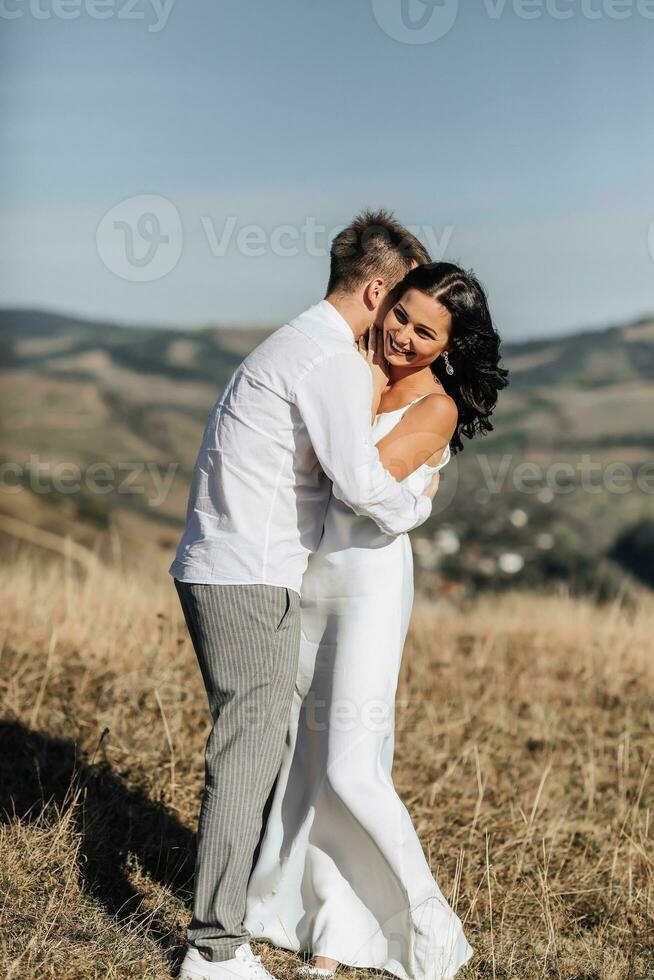 een elegant model- paar in de bergen in de zomer. een jong jongen en een meisje in een wit zijde jurk zijn wandelen Aan de helling tegen de achtergrond van de Woud en berg pieken. jongen kusjes een meisje foto