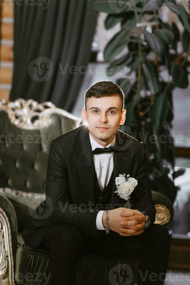 portret van positief jong bruidegom in zwart pak met wit overhemd glimlachen en zittend Aan comfortabel donker groen sofa in modern hotel kamer Aan bruiloft dag foto