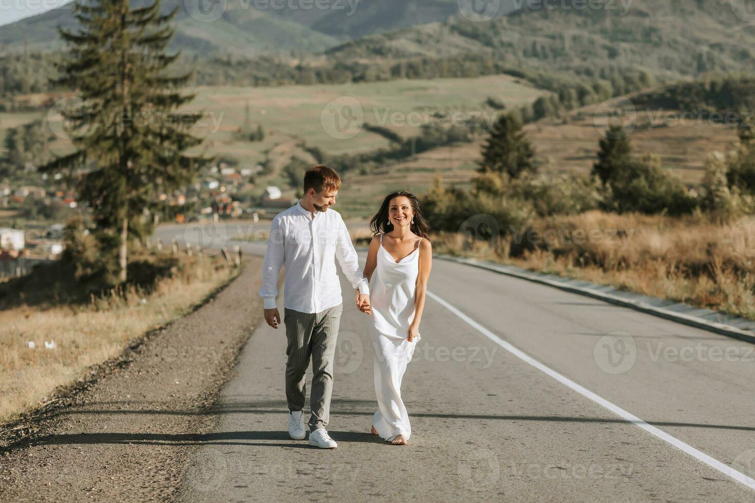 een elegant bruidegom in een wit overhemd en een schattig brunette bruid in een wit jurk zijn wandelen Aan een asfalt weg tegen de achtergrond van een Woud en bergen. bruiloft portret van pasgetrouwden. foto