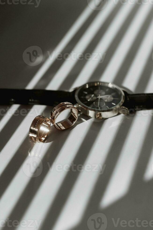 twee goud getextureerde bruiloft ringen Aan stralen van vallend licht in de buurt een polshorloge met een zwart leer band foto