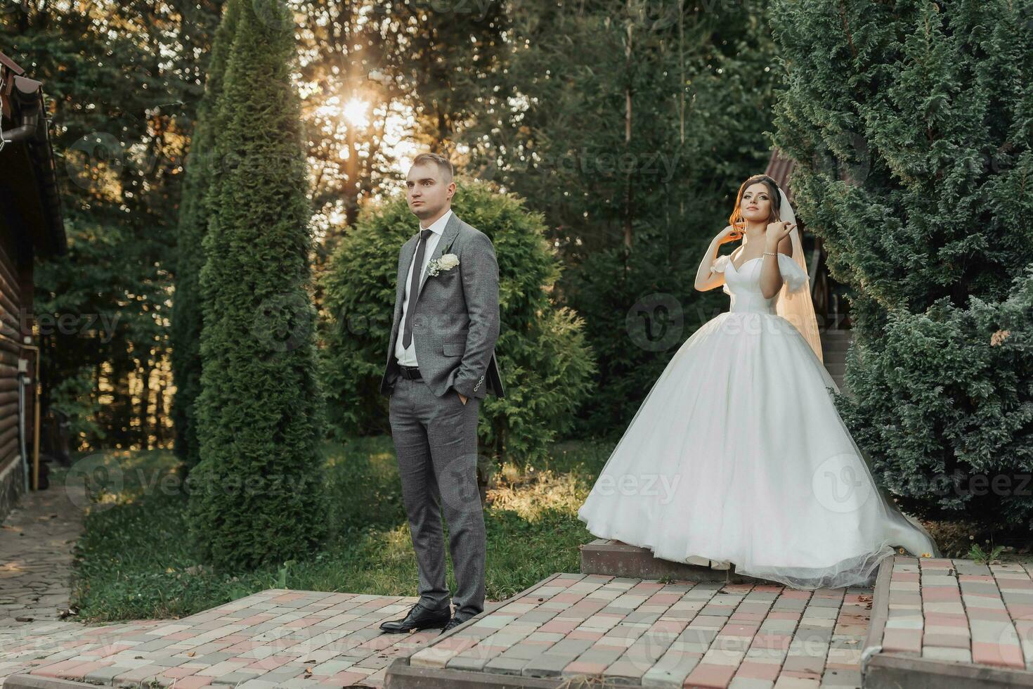 portret van de bruid en bruidegom in natuur. de bruidegom in een grijs pak is in de voorgrond, de bruid is wandelen achter hem, glimlachen in een wit omvangrijk jurk, Holding een boeket. elegant bruidegom foto