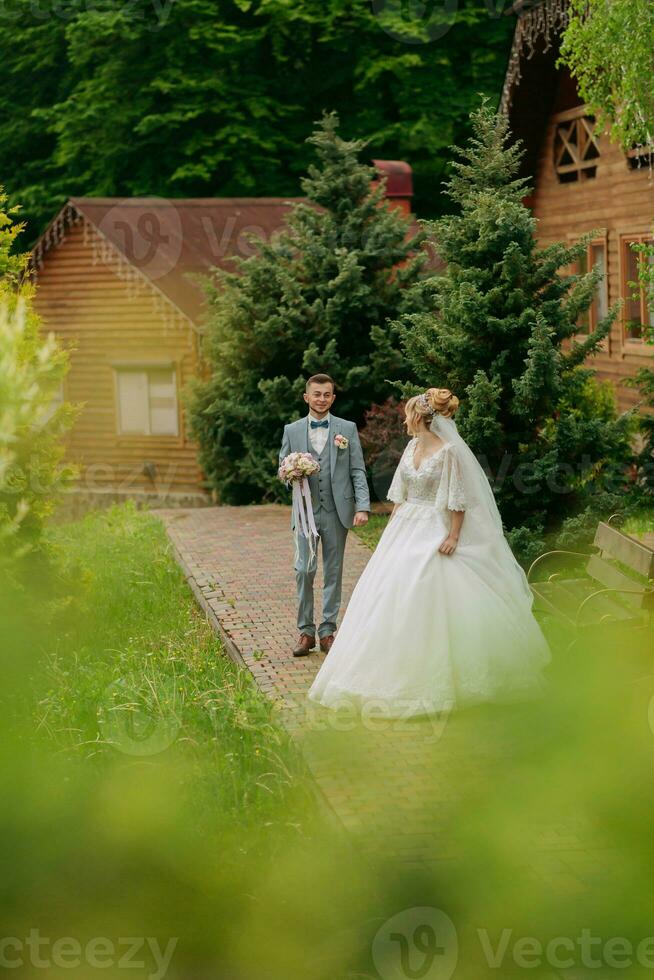 bruiloft in natuur. de bruid staat in voorkant van de bruidegom en looks over- zijn schouder tegen een achtergrond van bomen en een houten hut. elegant bruidegom. mooi bruid. foto