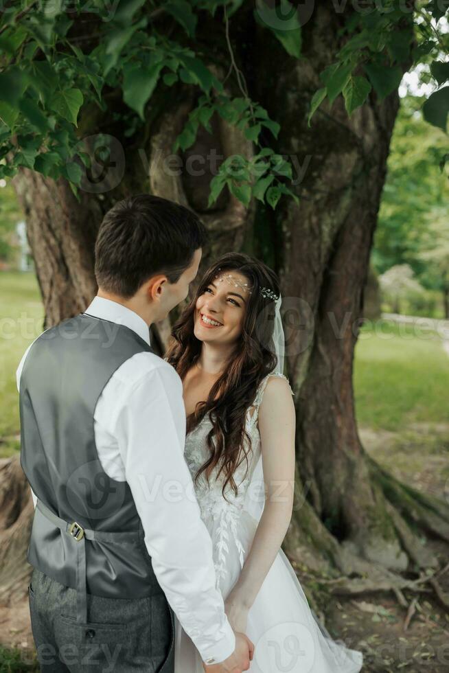 een mooi bruid met lang gekruld haar- in een elegant jurk looks Bij de bruidegom met liefhebbend ogen, glimlacht. portret van de bruid en bruidegom. voorjaar bruiloft. natuurlijk bedenken foto