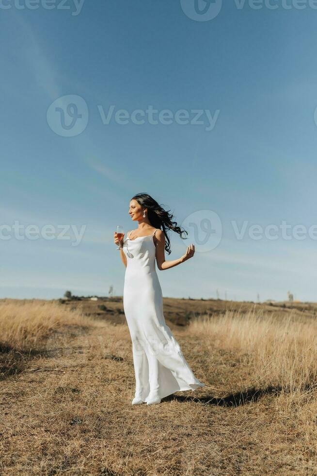 een meisje met lang, zwart, gekruld haar- blazen in de wind is staand gekleed in een wit zijde jurk, Holding een glas van Champagne in haar hand, tegen een achtergrond van blauw lucht. breed hoek foto