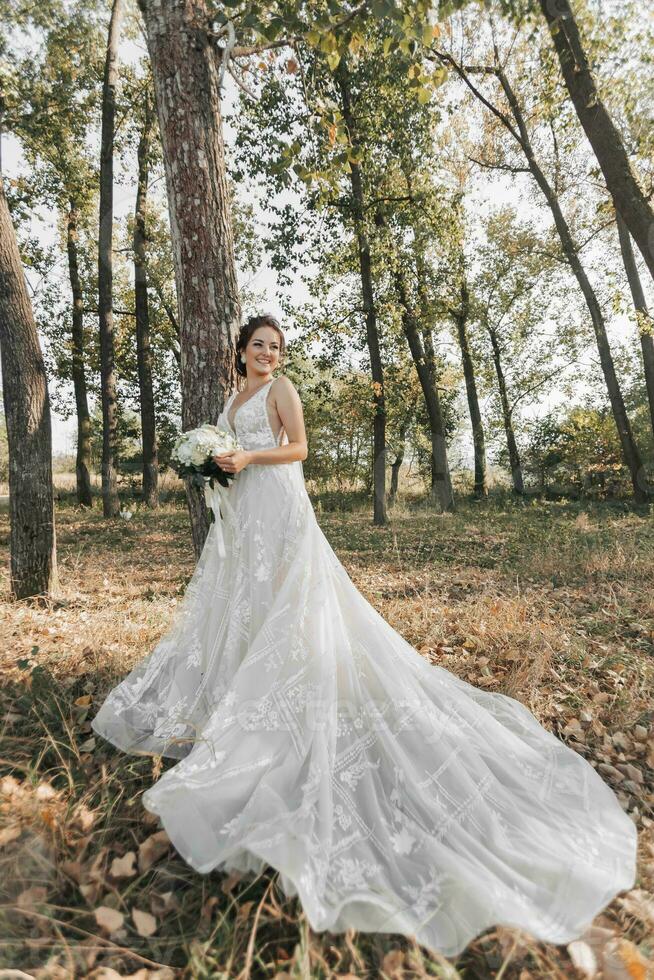 bruiloft foto in natuur. de bruid is staand in de Woud. de bruid in een mooi jurk met een lang trein, Holding haar boeket van wit rozen, glimlachen vriendelijke groet Bij de camera. portret