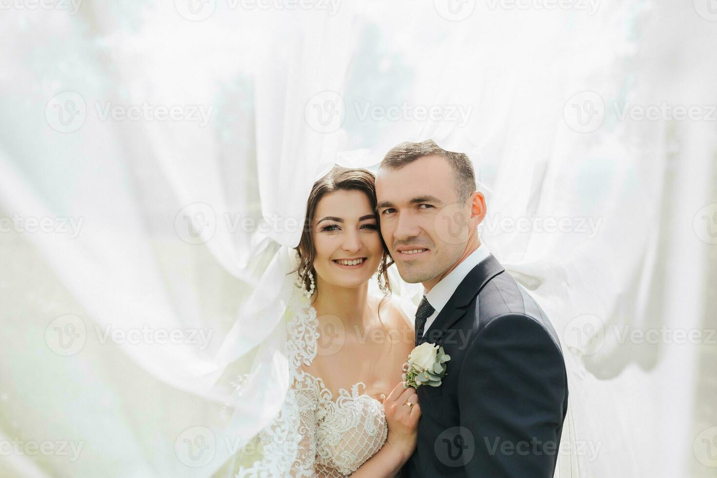 een krullend haar brunette bruid in een wit jurk en een gesluierd bruidegom omhelzing en kus. portret van de bruid en bruidegom. mooi bedenken en haar. bruiloft in natuur foto