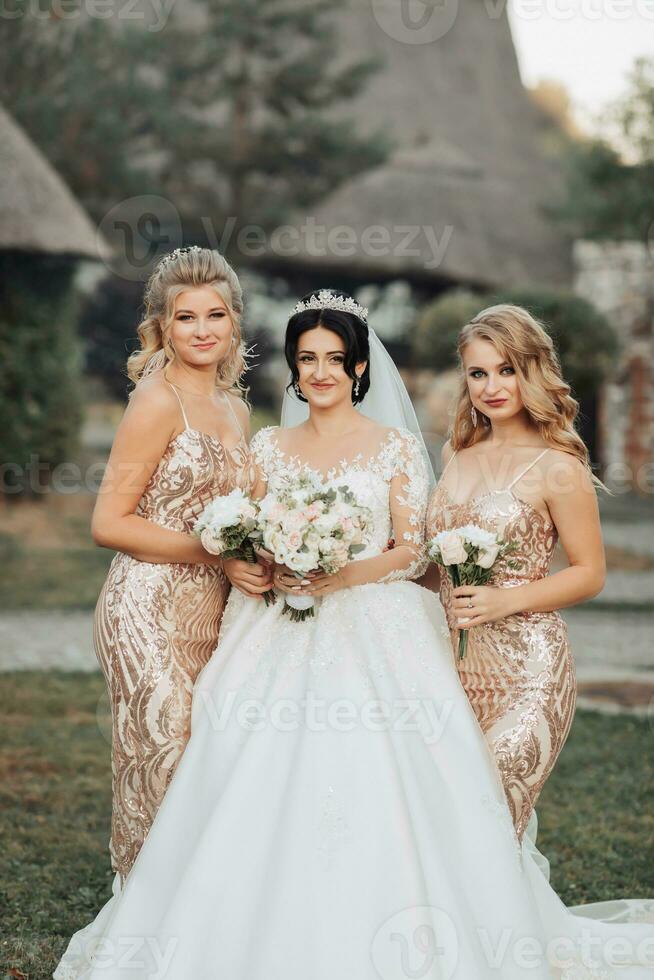 een brunette bruid in een wit elegant jurk met een kroon en haar blond vrienden in goud jurken houding met boeketten. bruiloft portret in natuur, bruiloft foto in een licht toon.