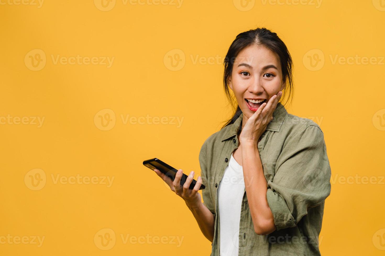 verraste jonge azië dame met behulp van mobiele telefoon met positieve uitdrukking, breed glimlachen, gekleed in casual kleding en camera kijken op gele achtergrond. gelukkige schattige blije vrouw verheugt zich over succes. foto
