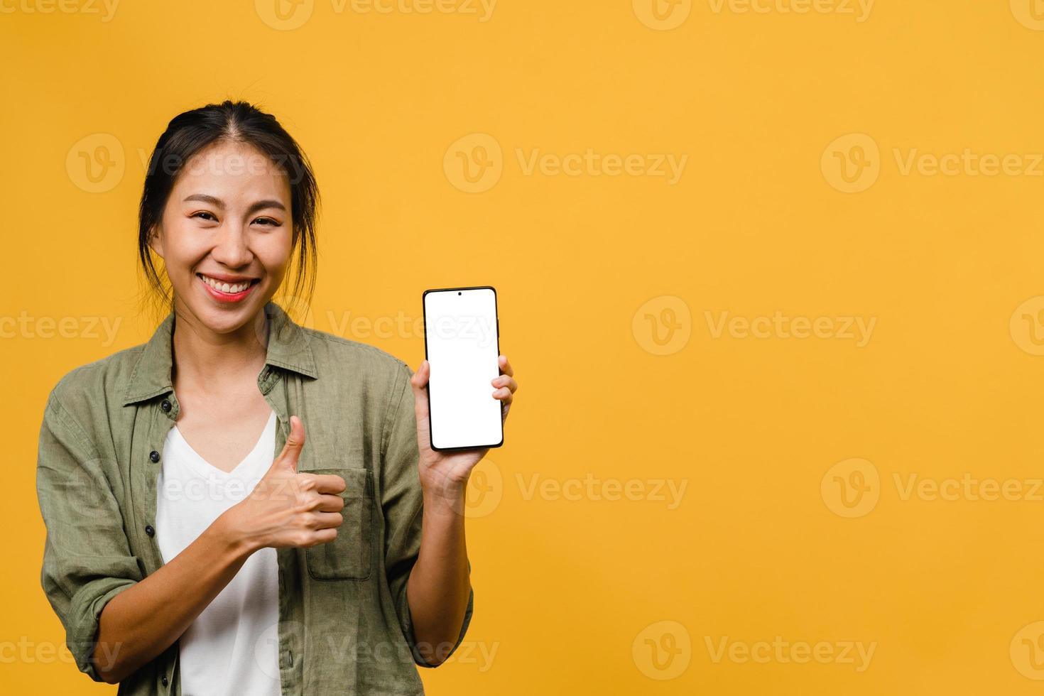 jonge aziatische dame toont een leeg smartphonescherm met positieve uitdrukking, glimlacht breed, gekleed in casual kleding en voelt zich gelukkig op gele achtergrond. mobiele telefoon met wit scherm in vrouwelijke hand. foto