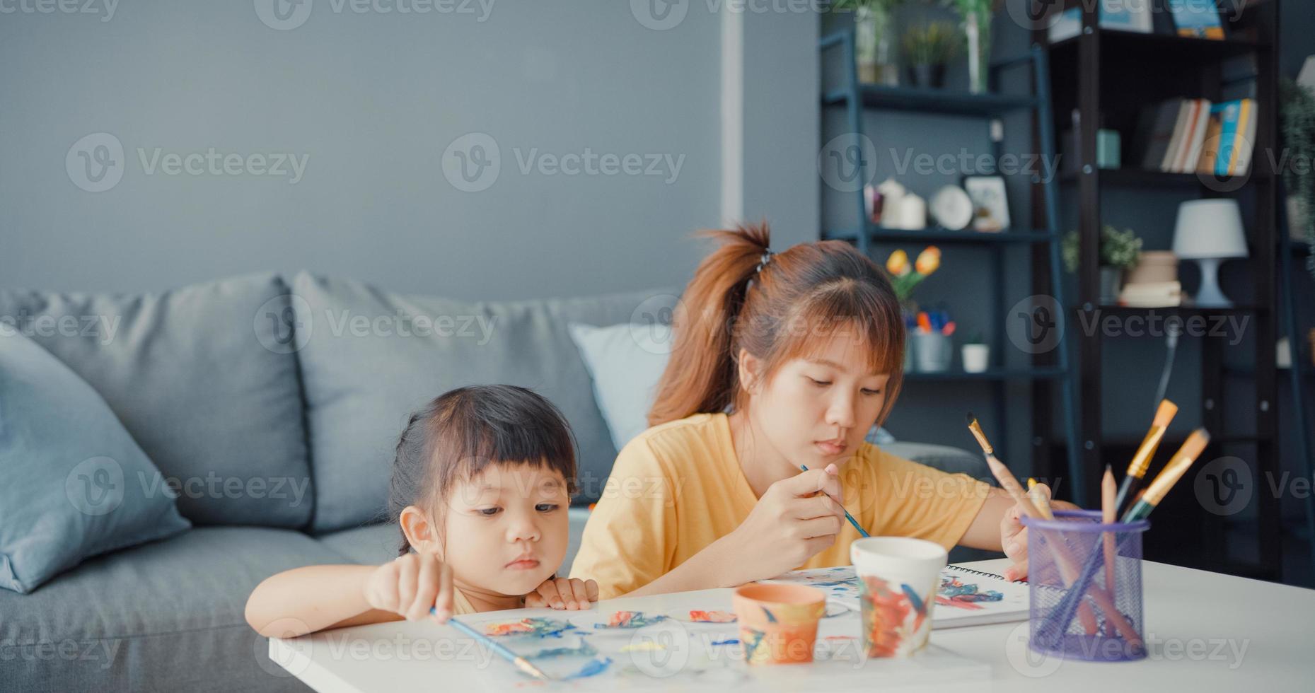 gelukkige vrolijke azië familie moeder leert peuter meisje verf keramische pot plezier ontspannen op tafel in de woonkamer thuis. tijd samen doorbrengen, sociale afstand, quarantaine voor coronaviruspreventie. foto