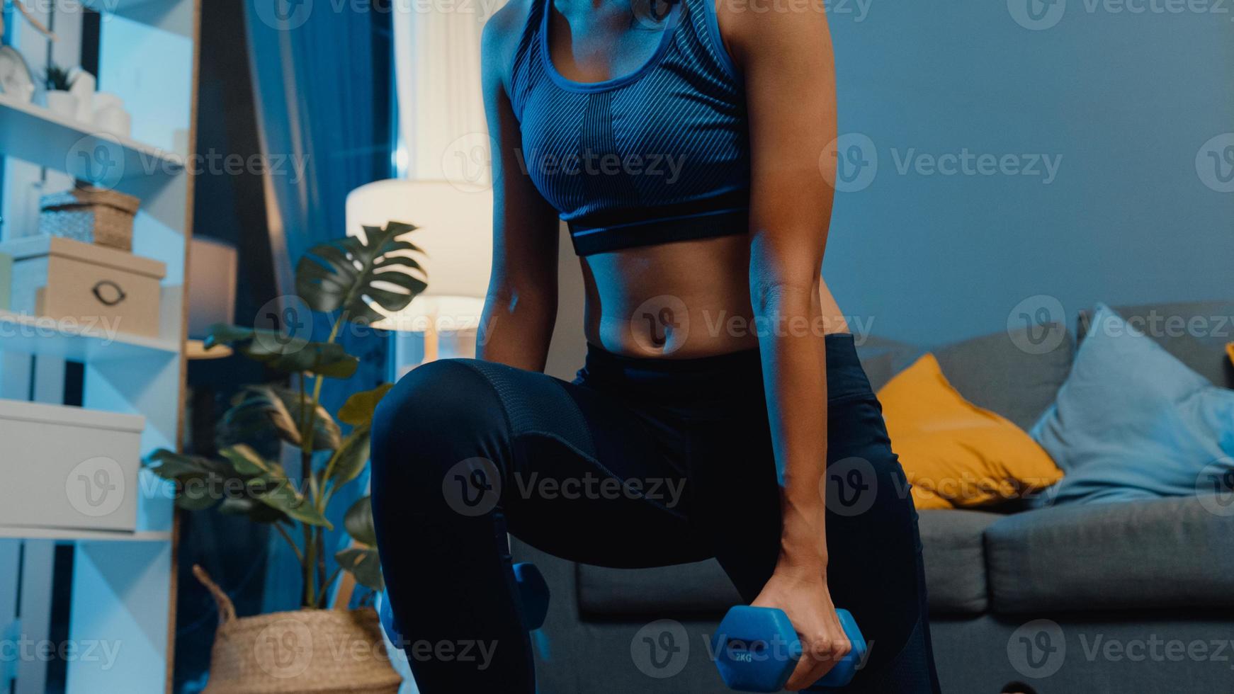 jonge Aziatische dame in sportkleding doet squat met halters in uitgestrekte armen oefening uit te werken in de woonkamer thuis 's nachts. sport- en recreatieactiviteit, quarantaine voor coronavirusconcept. foto