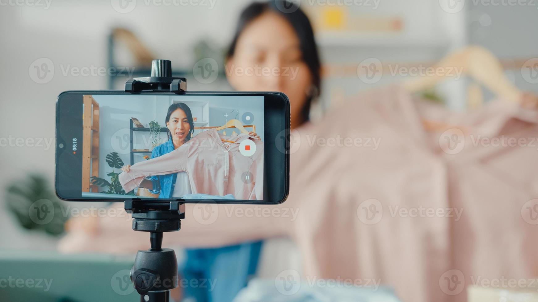 jonge aziatische dame modeontwerper met behulp van mobiele telefoon die inkooporder ontvangt en kleding laat zien die video livestreaming online opneemt in de winkel. eigenaar van een klein bedrijf, online marktleveringsconcept. foto