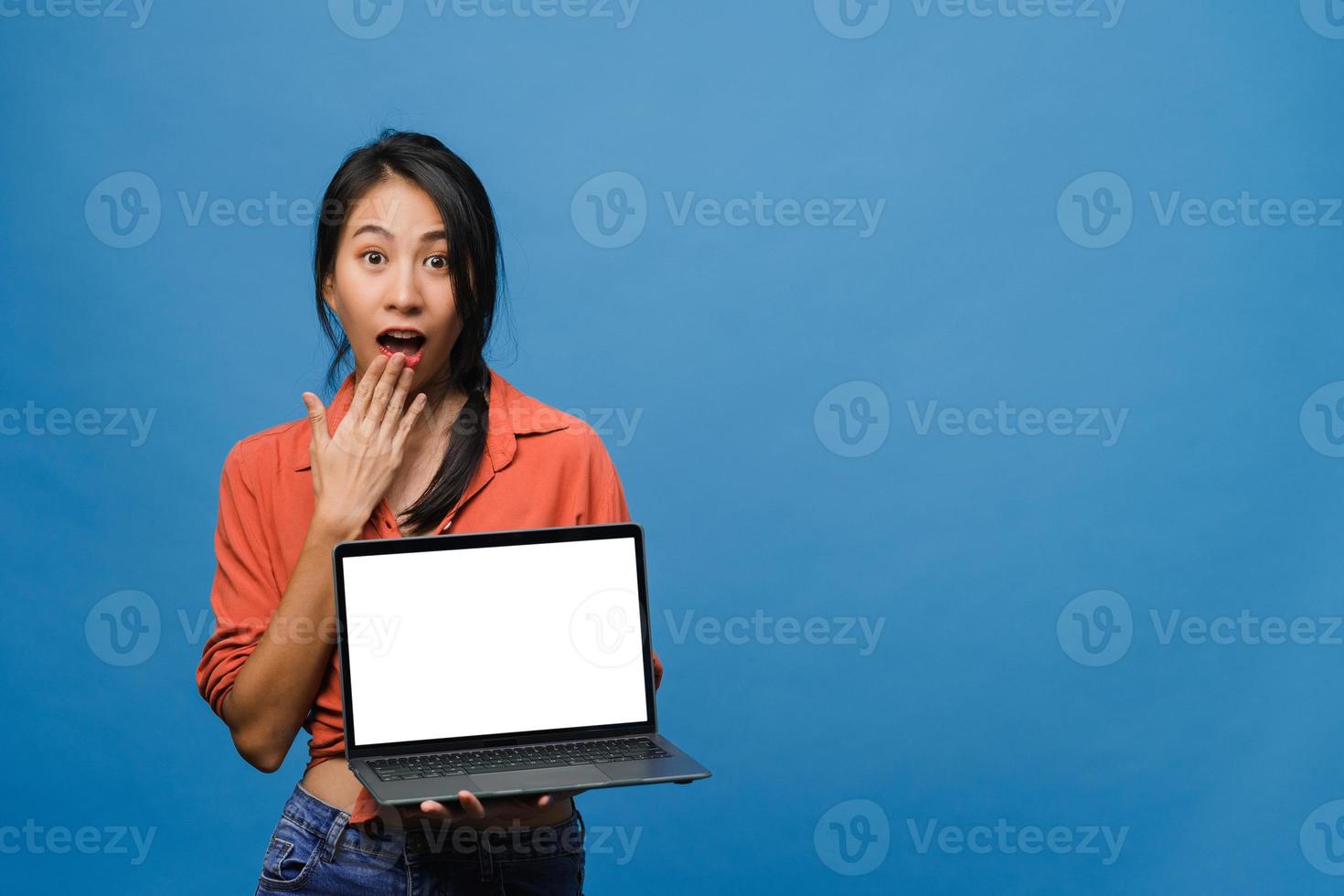 jonge aziatische dame toont een leeg laptopscherm met positieve uitdrukking, glimlacht breed, gekleed in casual kleding die geluk voelt geïsoleerd op een blauwe achtergrond. computer met wit scherm in vrouwelijke hand. foto