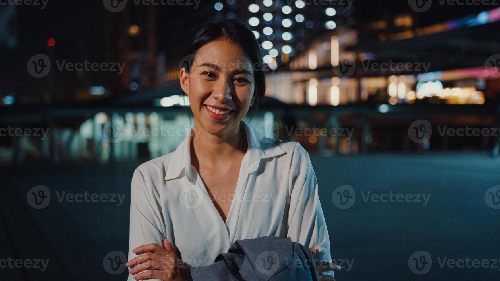succesvolle jonge Aziatische zakenvrouw in mode kantoorkleding glimlachend en kijkend naar de camera terwijl ze 's nachts gelukkig alleen staat in de stedelijke moderne stad. bedrijf onderweg en forenzenconcept. foto