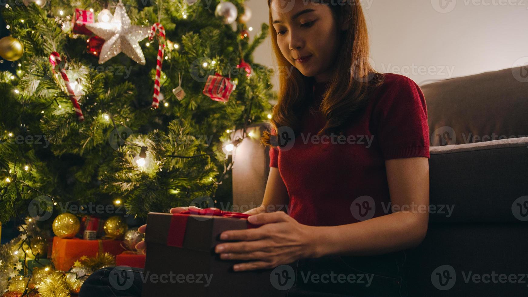 jonge azië vrouw plezier openen x'mas huidige doos in de buurt van kerstboom versierd met ornamenten in de woonkamer thuis. prettige kerstnacht en gelukkig nieuwjaar vakantie festival. foto