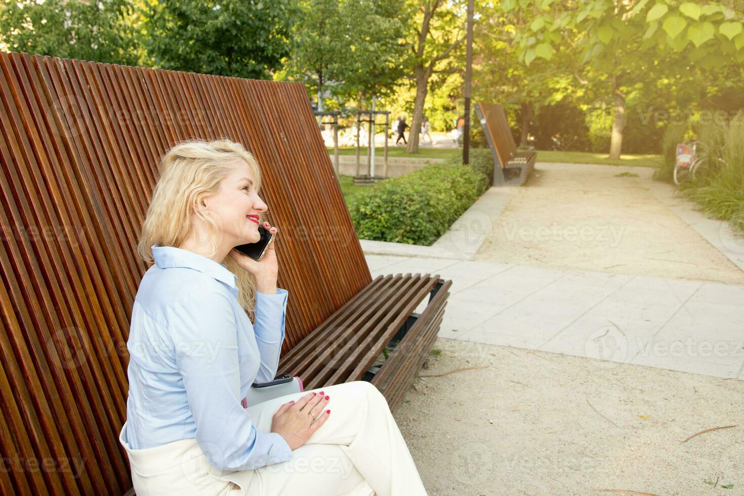 een mooi hoor jong vrouw is zittend Aan een bank in de park, pratend Aan de telefoon, video communicatie. zomer dag. kant visie foto