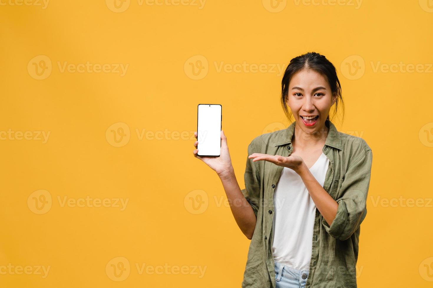 jonge aziatische dame toont een leeg smartphonescherm met positieve uitdrukking, glimlacht breed, gekleed in casual kleding en voelt zich gelukkig op gele achtergrond. mobiele telefoon met wit scherm in vrouwelijke hand. foto