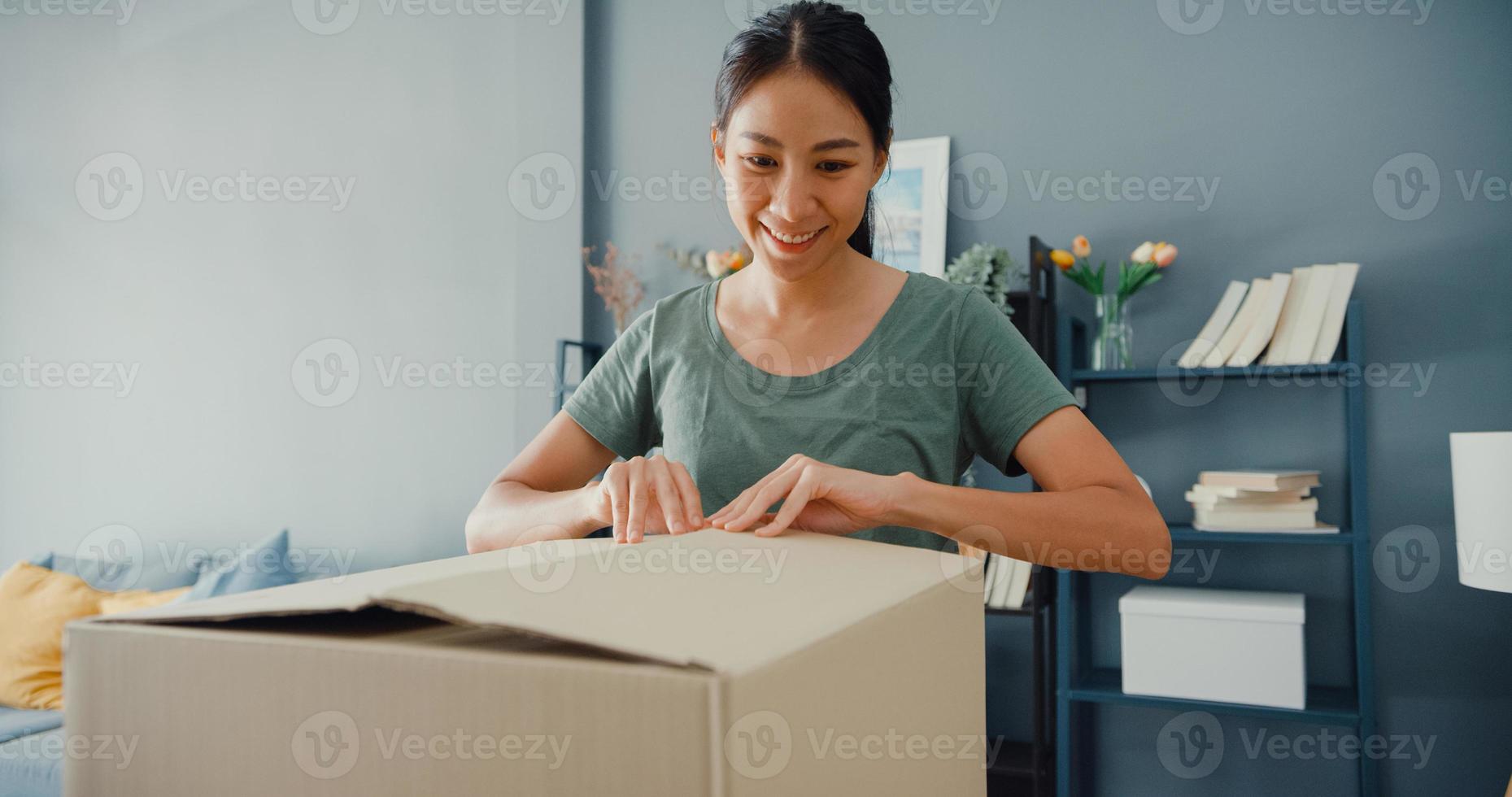 gelukkige mooie azië dame unboxing kartonnen leveringspakket van online marktplaats in de woonkamer bij huis. tevreden shopper in internet unboxing goederen, online winkelen en levering concept. foto