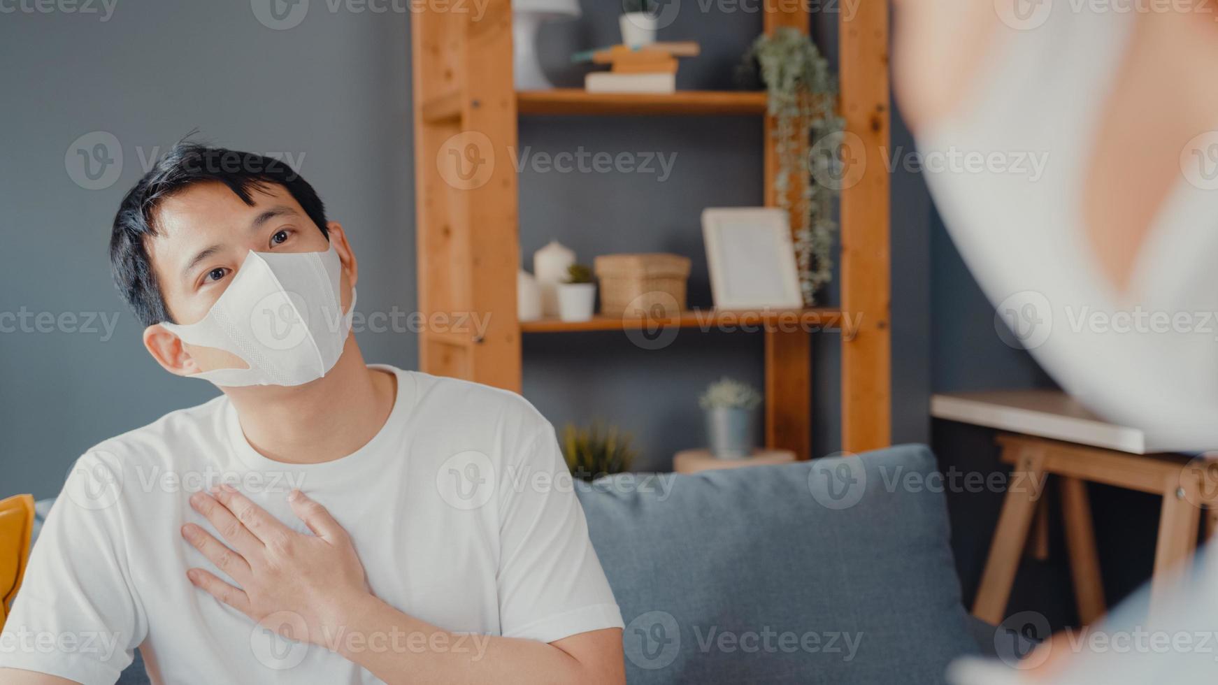 jonge azië vrouwelijke arts arts draagt gezichtsmasker met behulp van digitale tablet die goed nieuws over gezondheidstests deelt met gelukkige mannelijke patiënt die in huis op de bank zit. medische verzekering, bezoek patiënt thuis concept. foto
