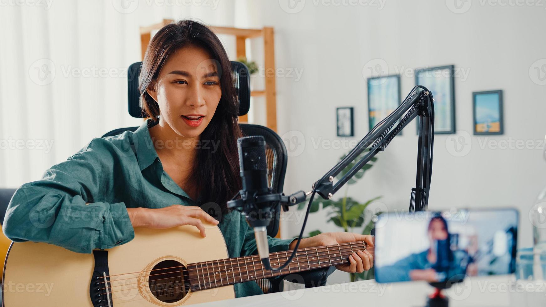 tiener azië meisje influencer gitaar spelen muziek gebruik microfoon record met smartphone voor online publiek luister thuis. vrouwelijke podcaster maakt audiopodcast vanuit haar thuisstudio, blijf thuis concept. foto