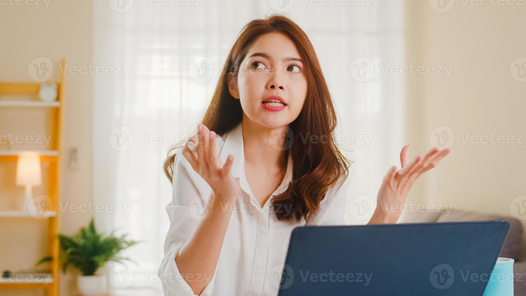 Aziatische zakenvrouw die laptop gebruikt, praat met collega's over het plan in een videogesprek terwijl ze slim vanuit huis in de woonkamer werkt. zelfisolatie, sociale afstand, quarantaine voor coronaviruspreventie. foto