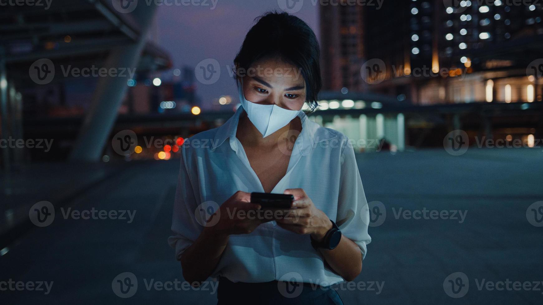 jonge azië zakenvrouw in mode kleding met gezichtsmasker met behulp van smartphone sms-bericht te typen terwijl je 's nachts buiten in de stedelijke stad staat. sociale afstand om verspreiding van het covid-19-concept te voorkomen. foto